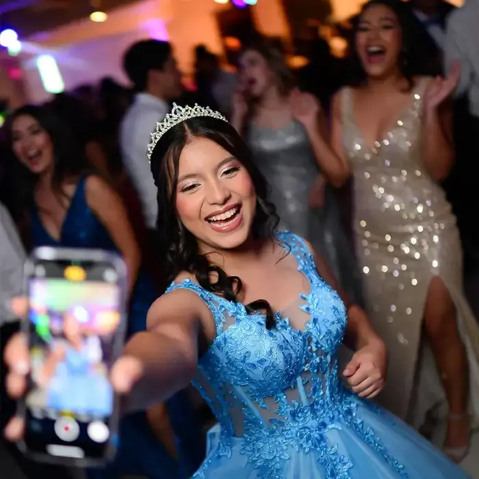Quinceañera con vestido azul y tiara ríe en la pista mientras un invitado la graba con el teléfono.