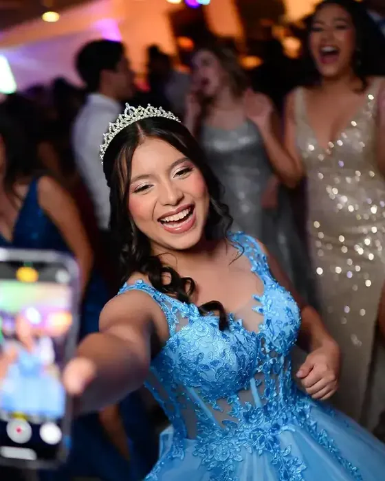 Quinceañera con vestido azul y tiara ríe en la pista mientras un invitado la graba con el teléfono.